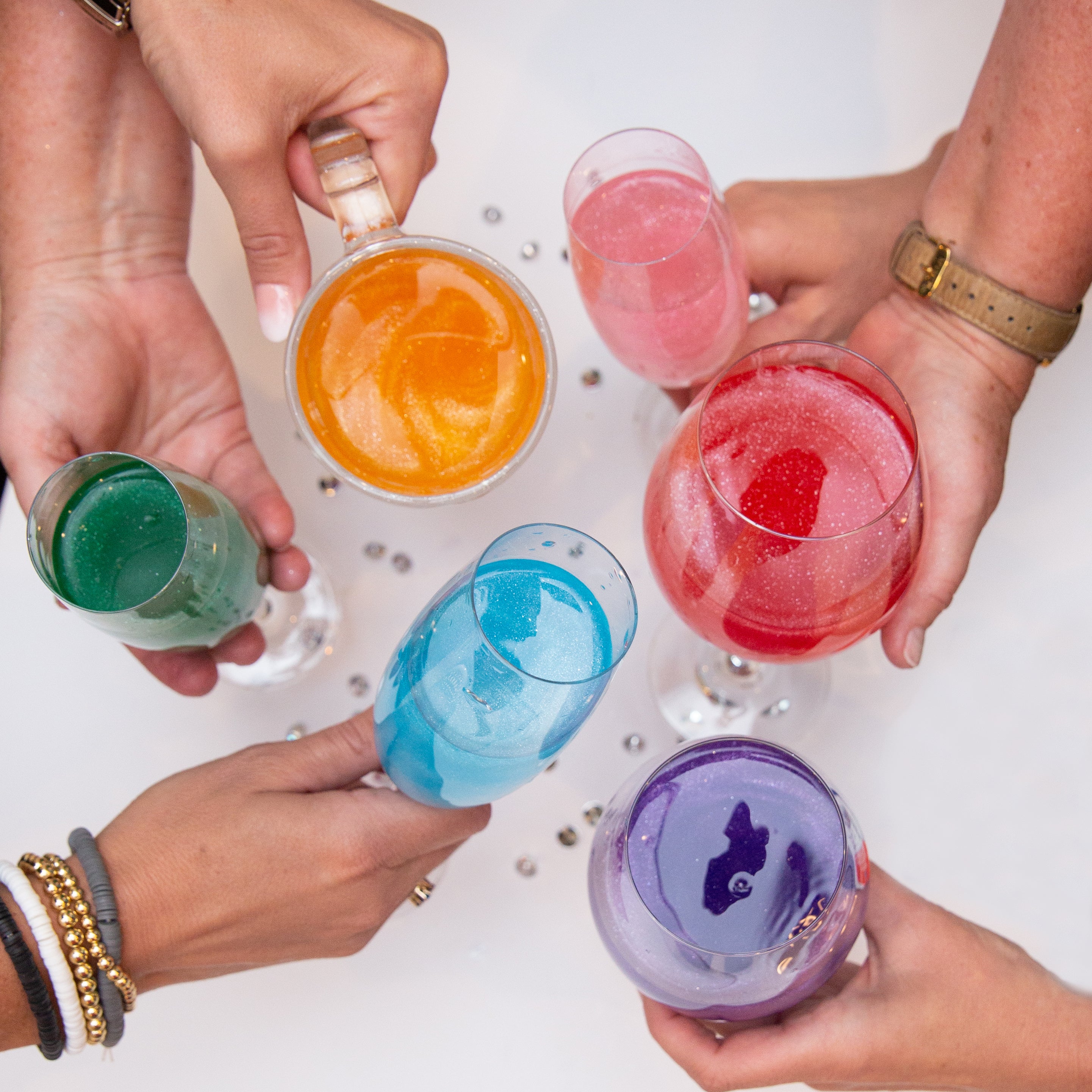 Six women clinking their glasses together to "cheers". Each glass has a different color of Sugar Mama Shimmer. Cha Cha Red, Violet Vibes, Blue La La, Mango Mama and Glow Up green in champagne and wine glasses.